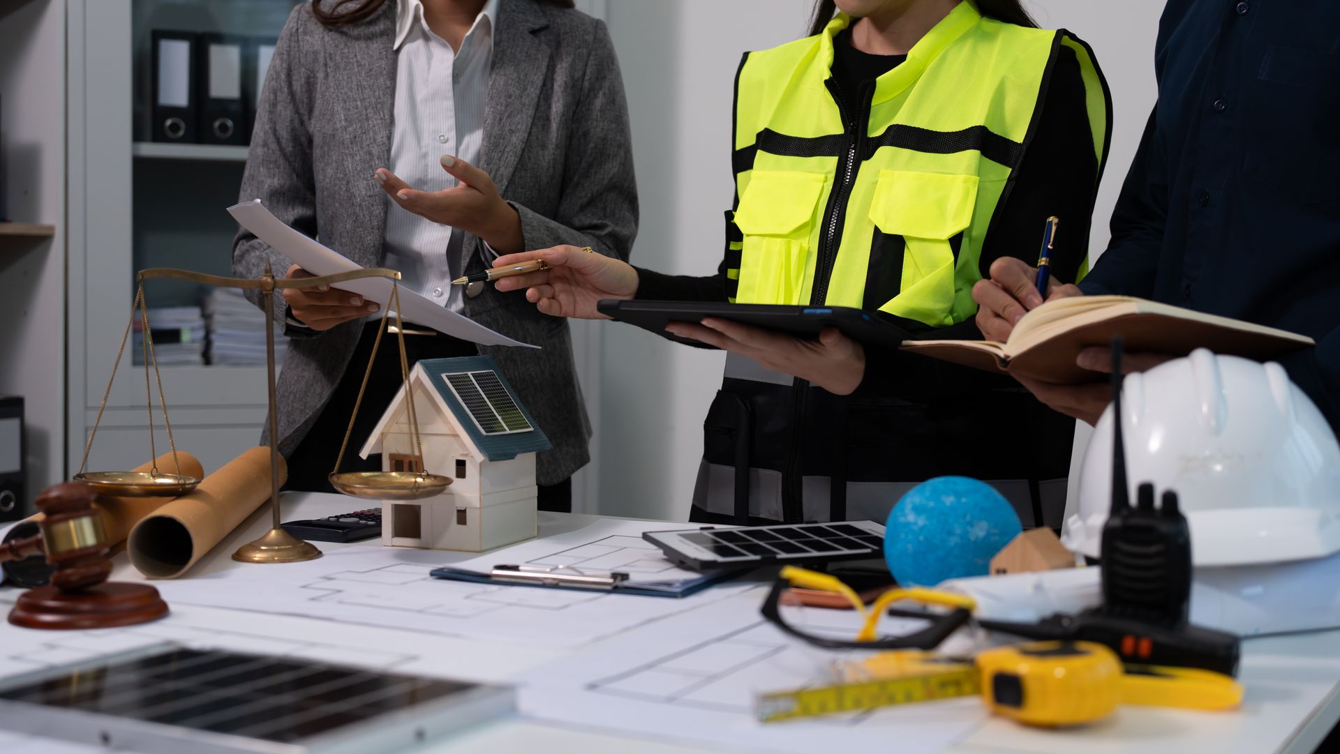 Two people in workwear reviewing blueprints at a cluttered construction desk with tools and a hard hat
