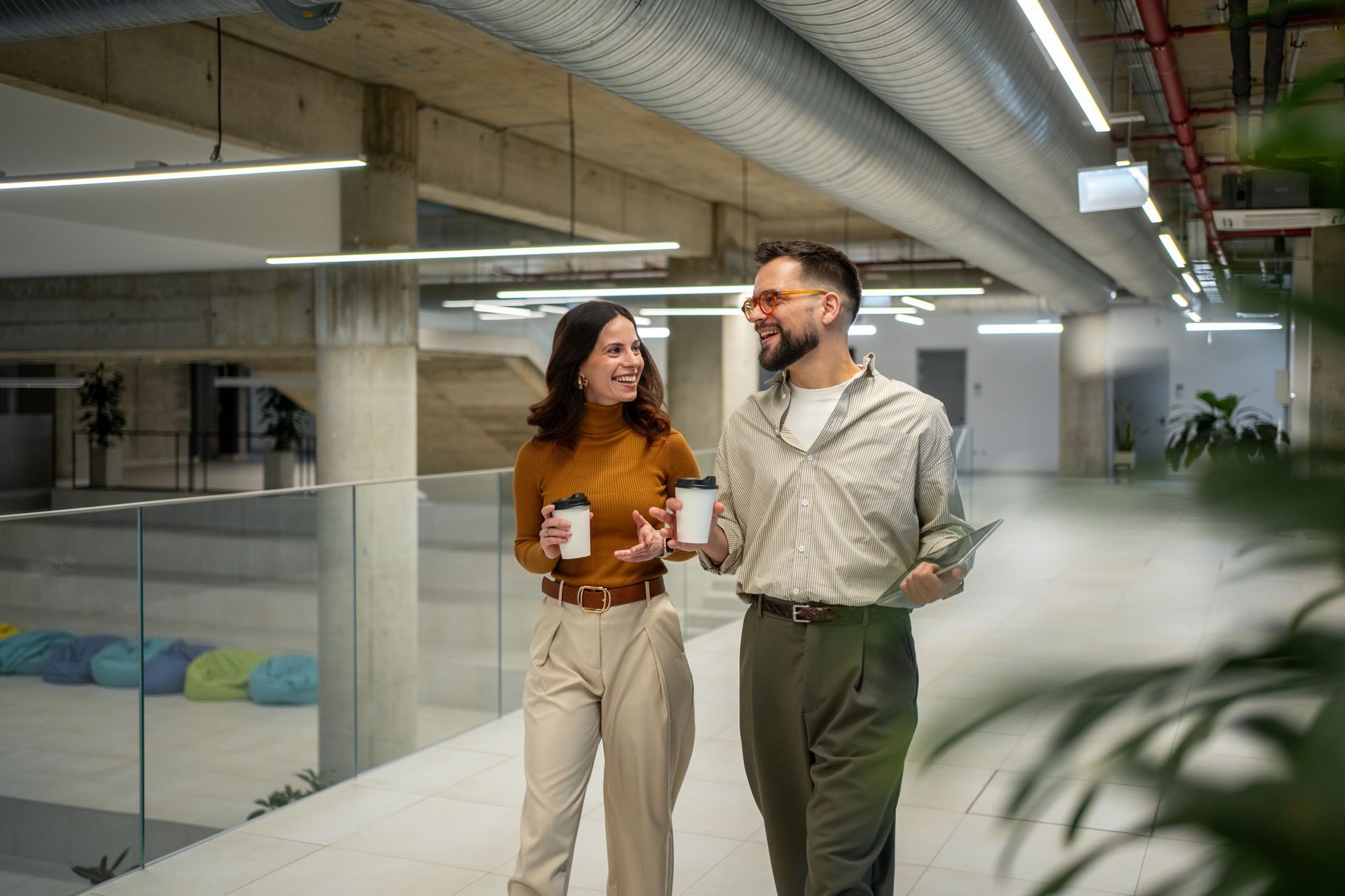 Two coworkers walking and talking in a modern office hallway, holding drinks and smiling