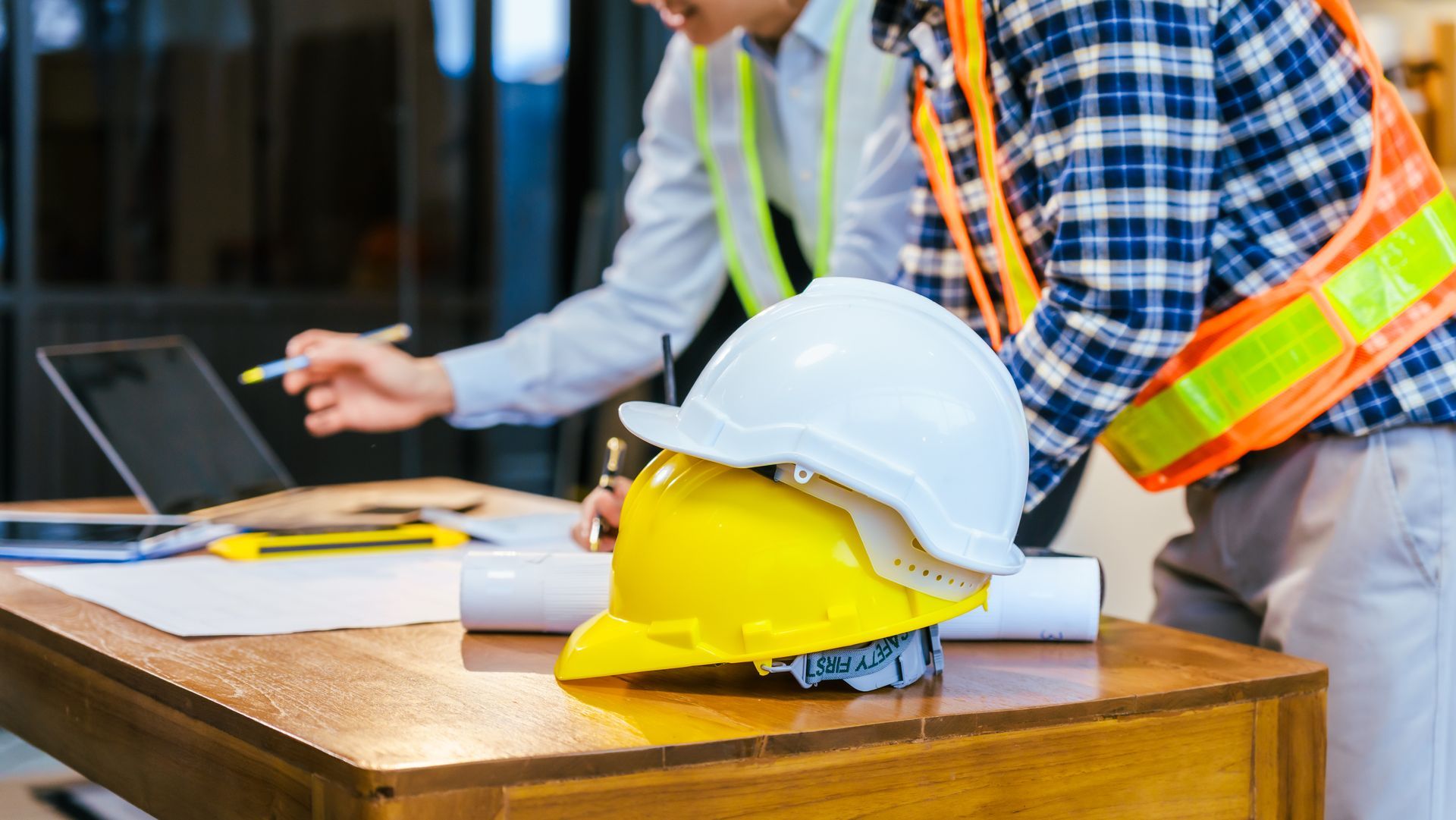 Construction workers in safety vests and hard hats reviewing plans at a table with a laptop