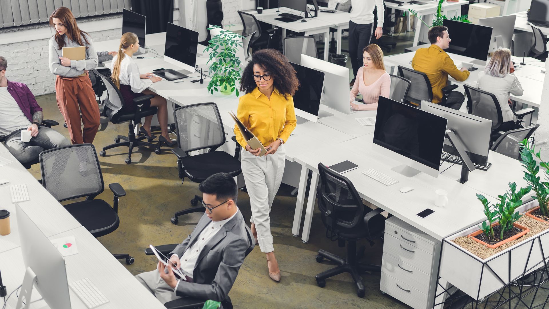 Open-plan office with people working at desks and computers, including one person in a yellow shirt standing.