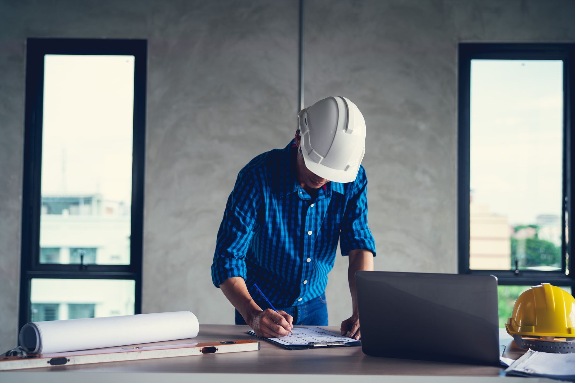 Construction worker in white hard hat writing plans at desk with laptop, blueprint, and yellow helmet
