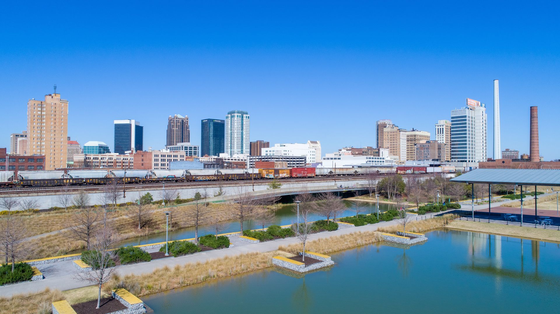 City skyline with modern high-rises and a waterfront park under a clear blue sky.