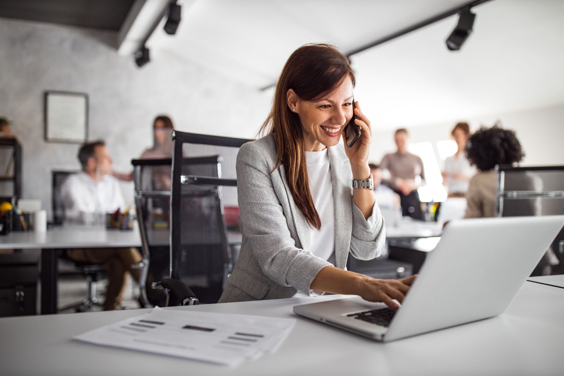 Woman working on a laptop at a desk in a bright office, smiling and on the phone Woman working on a laptop at a desk in a bright office, smiling and on the phone