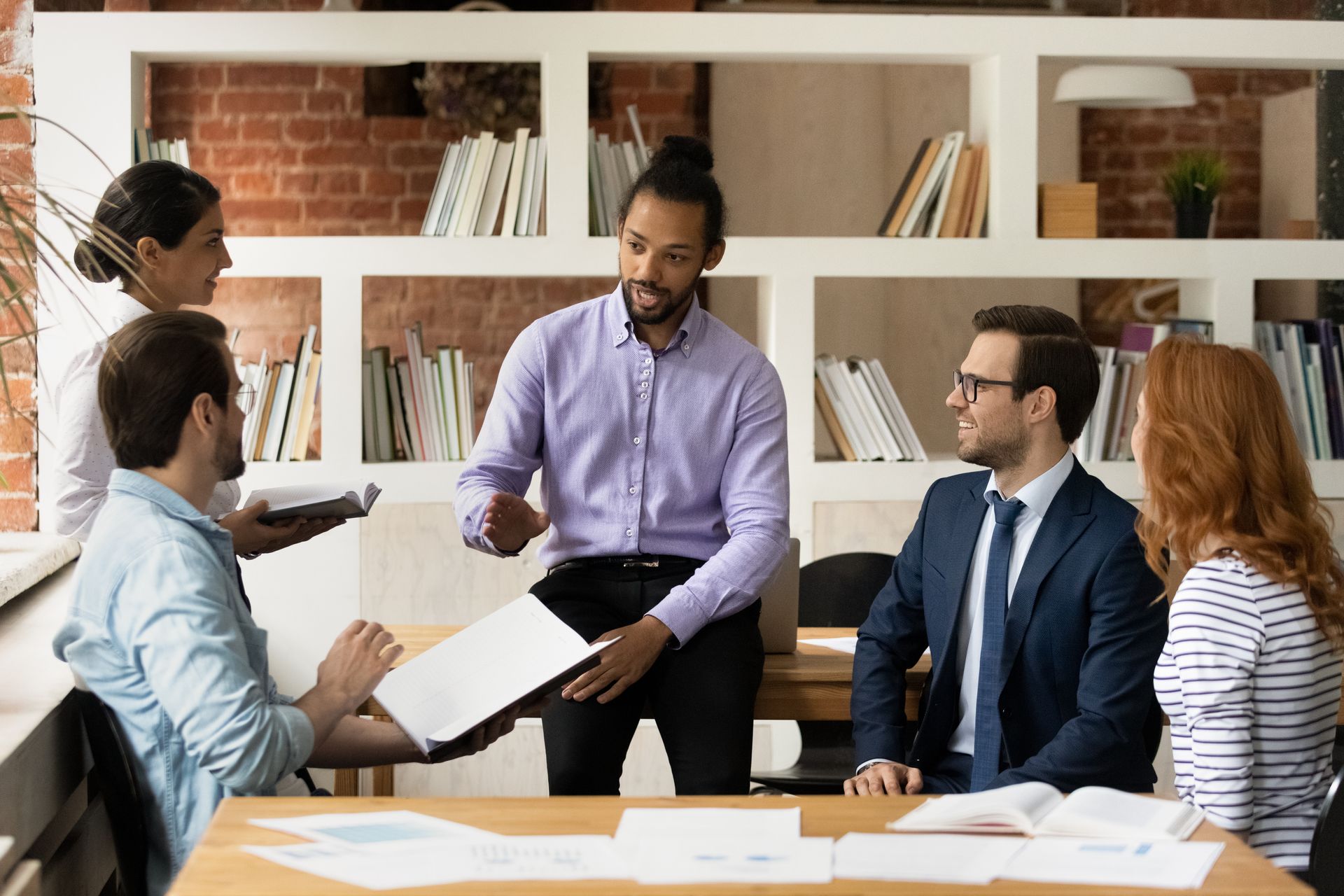 Team meeting in a bright office, with five people discussing documents around a table.