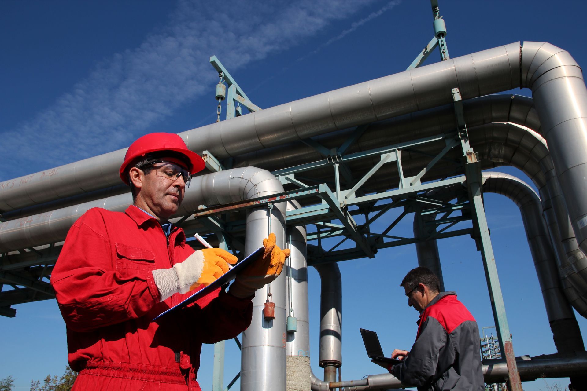 Two workers in red and dark uniforms inspect industrial pipes outdoors under a blue sky.