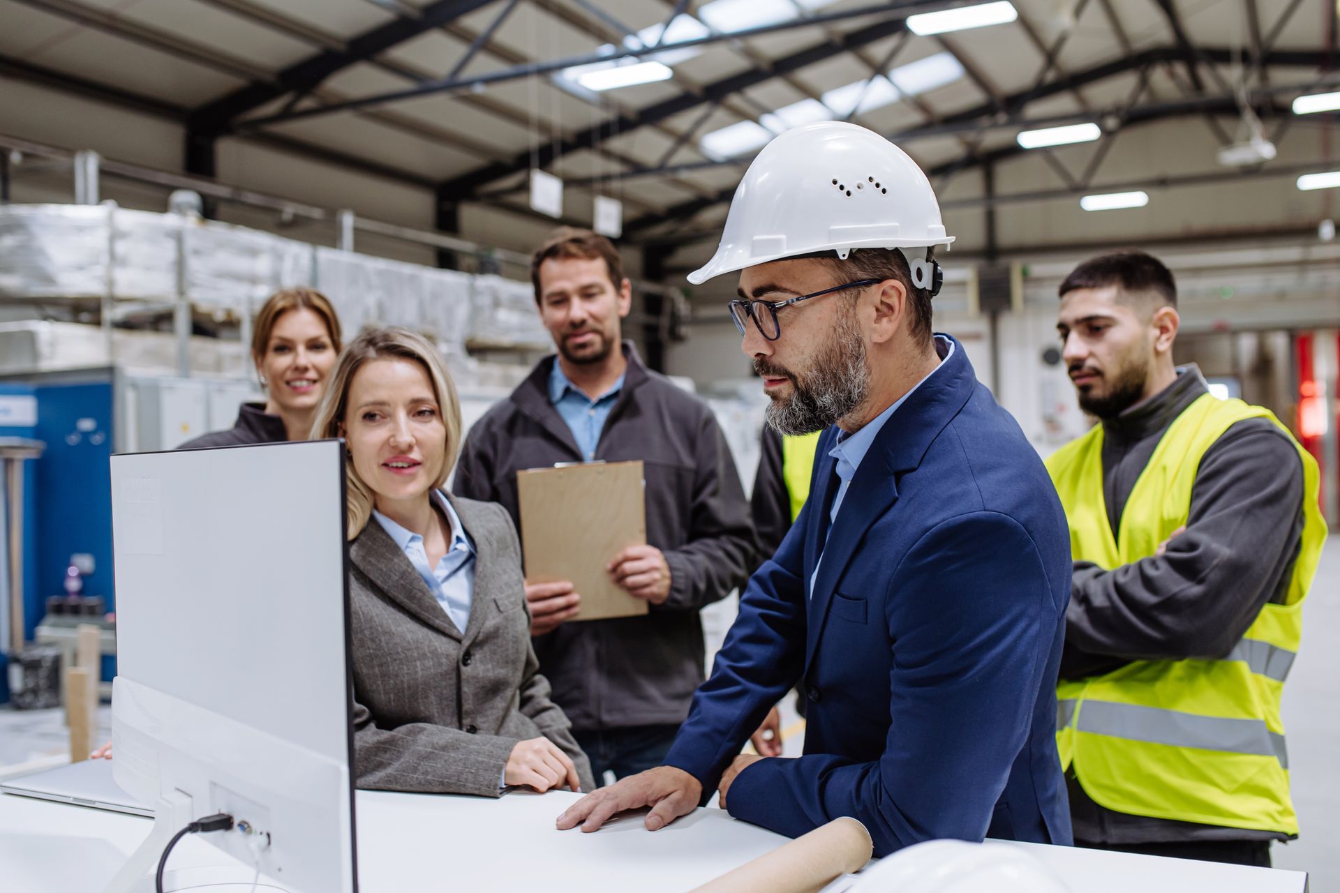 Team of workers in a factory meeting around a computer, with a man in a hard hat and safety vest beside them