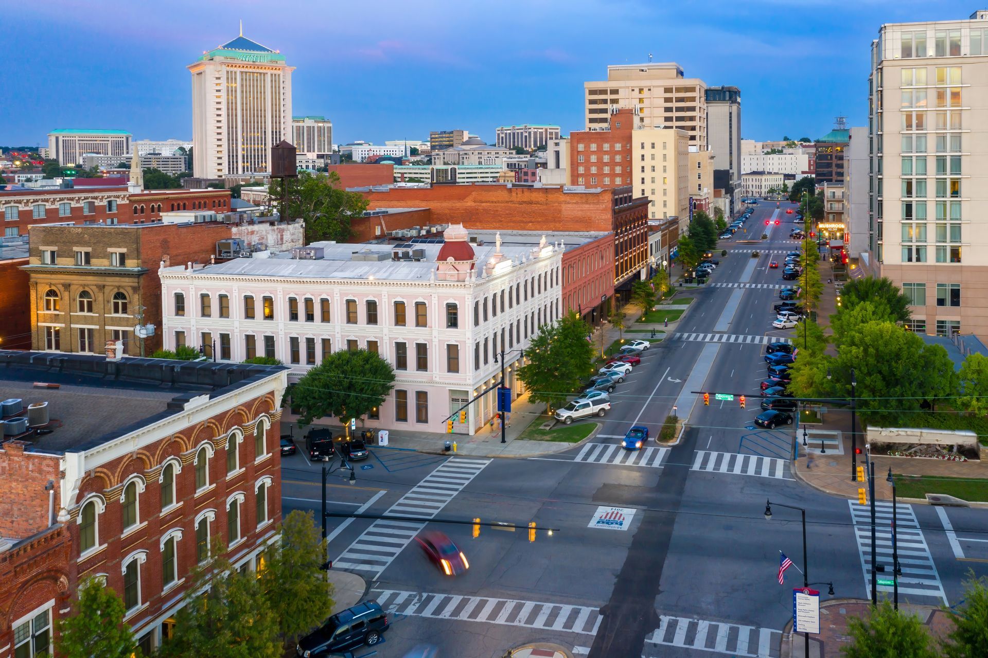 Downtown city street with crosswalks, brick and white buildings, and tall towers at dusk