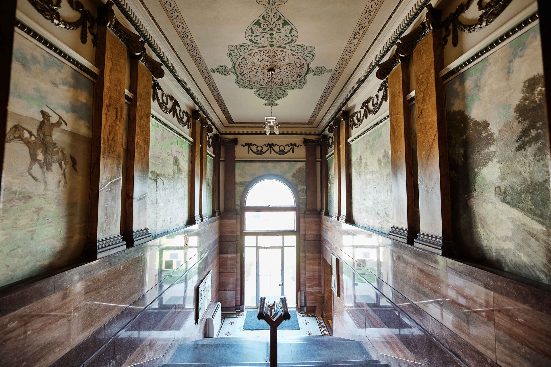 Ornate hallway with murals, a decorative ceiling, and a large arched doorway leading outside.