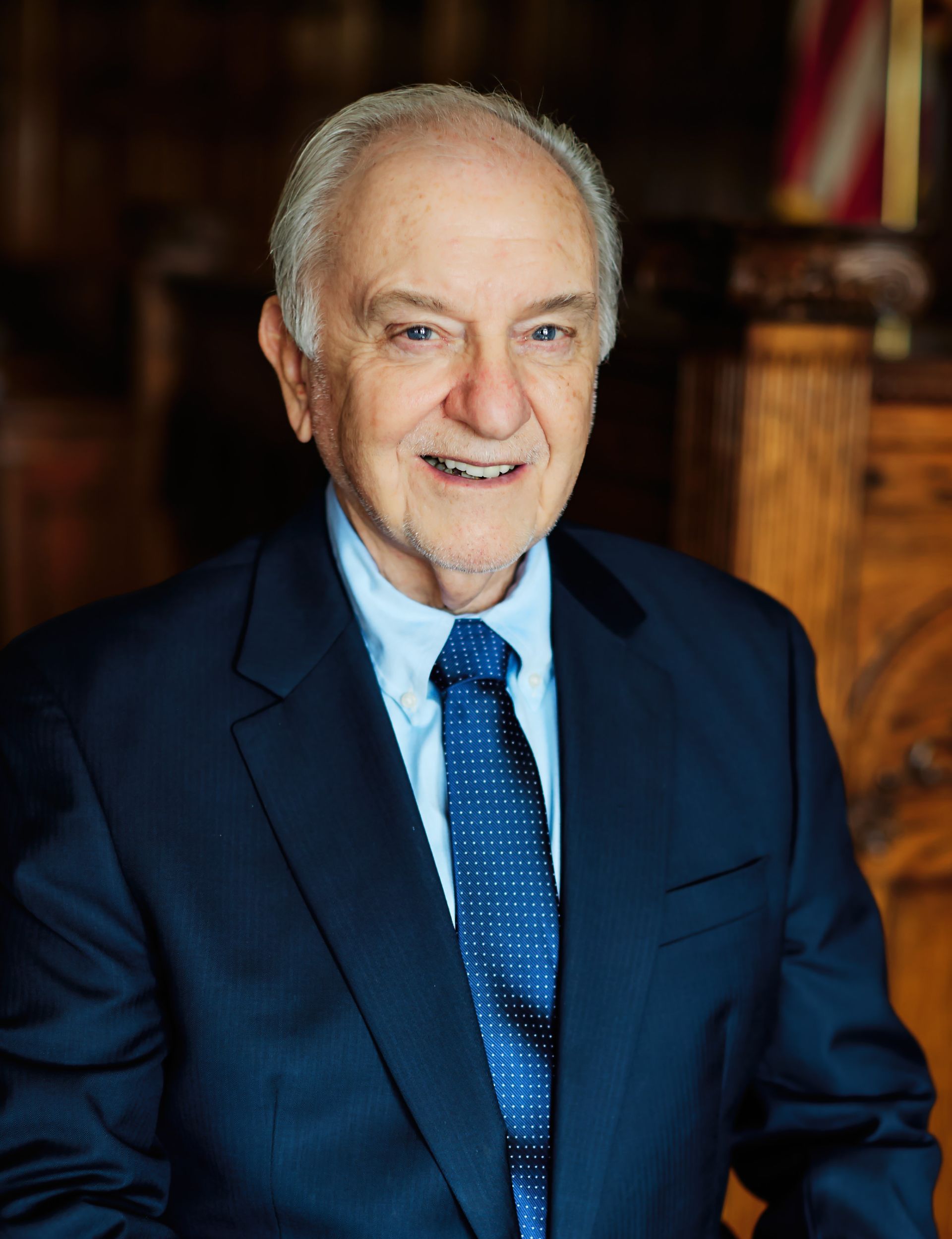 Smiling man in a navy suit and blue tie, standing indoors against a warm, blurred background