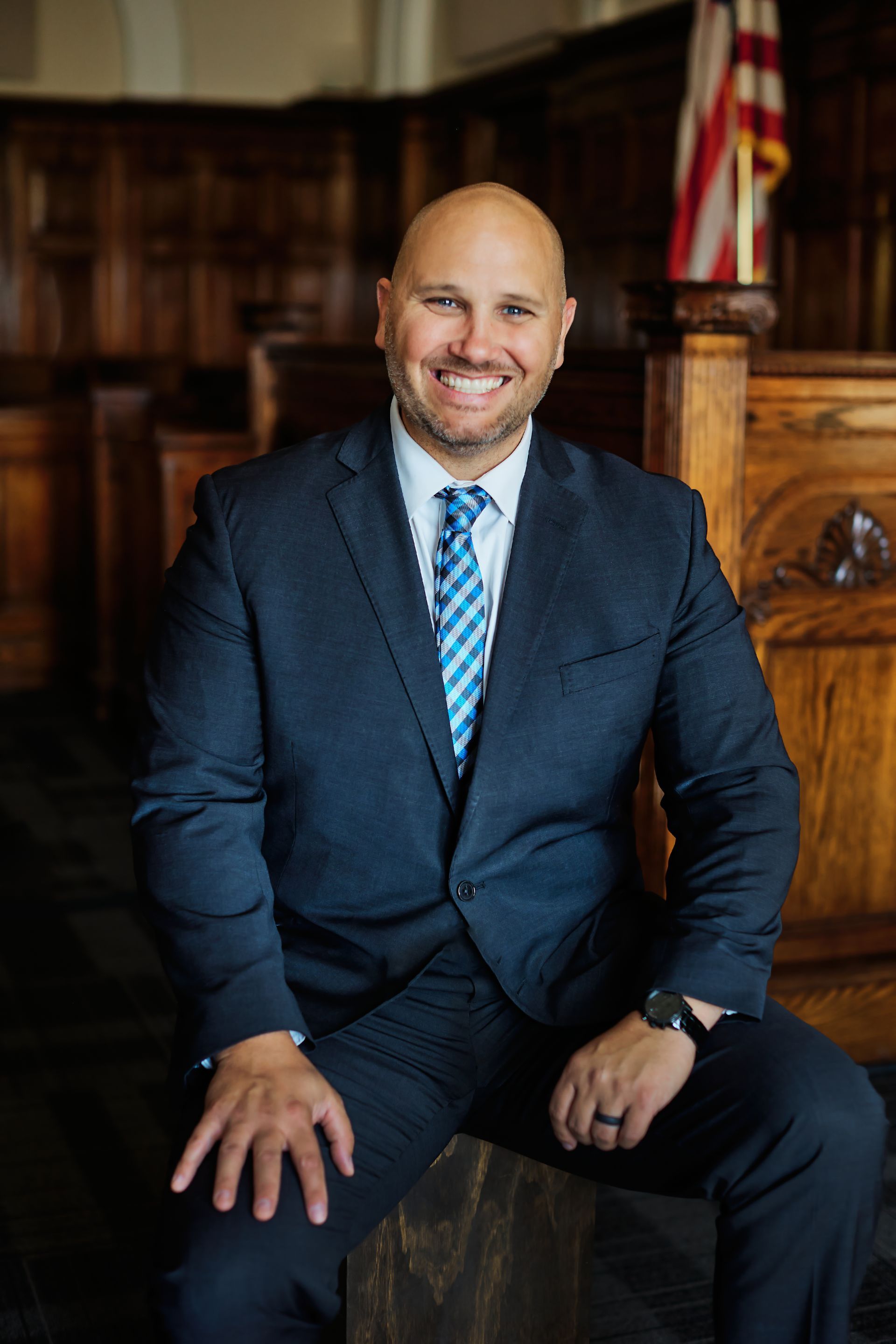 Man in a navy suit with a blue striped tie seated in a wooden courtroom, smiling at the camera.