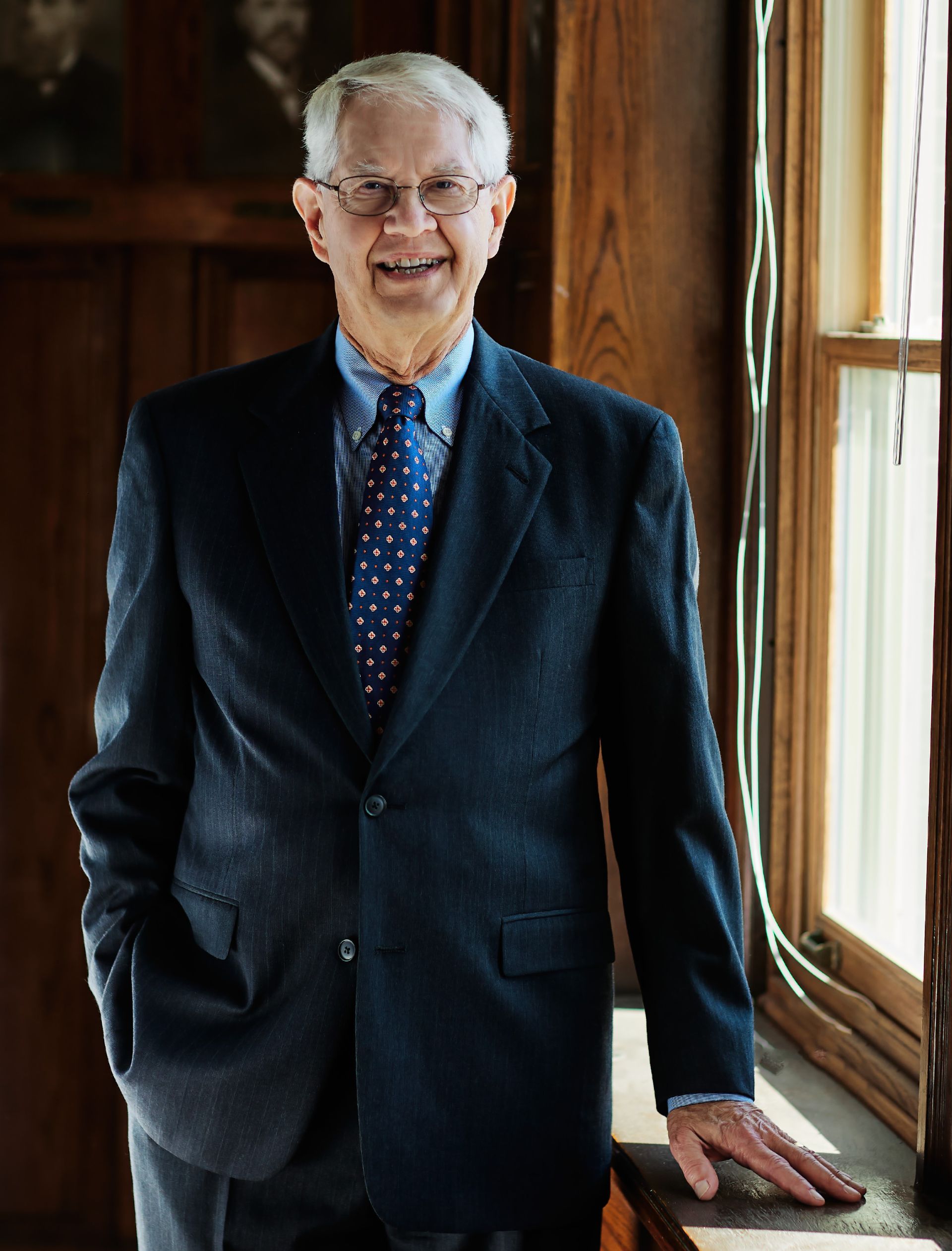 Smiling man in a dark suit standing by a sunlit window indoors.