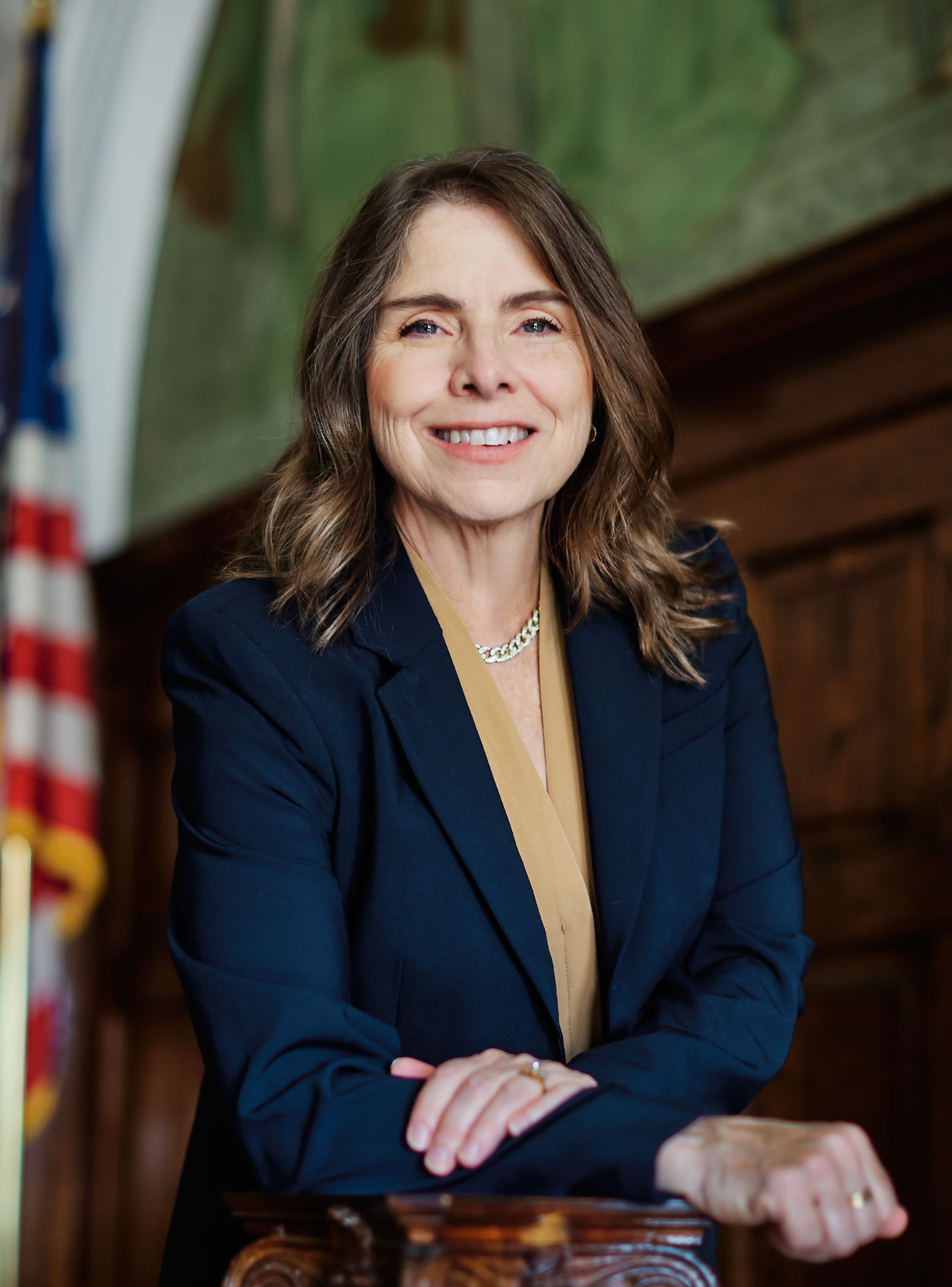 Woman in a navy blazer, smiling and leaning on a desk in a formal office setting.