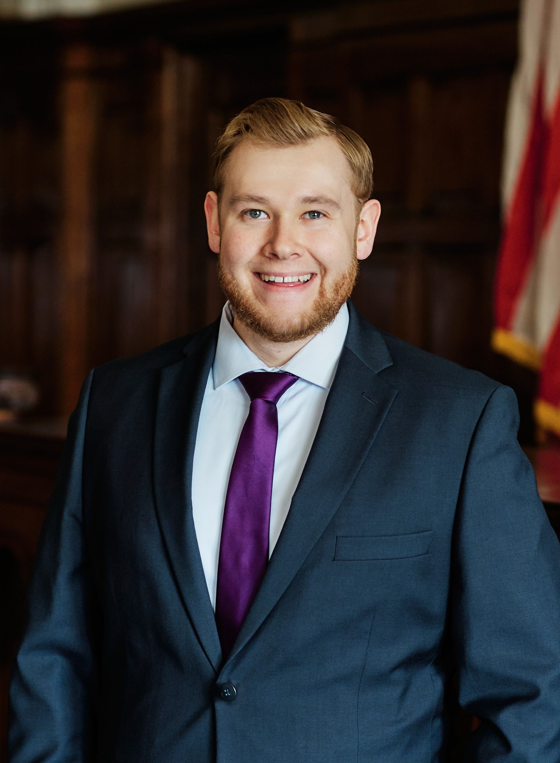 Smiling person in a navy suit and purple tie standing indoors, with wood paneling and a flag in the background