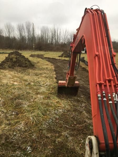Orange excavator arm digging a muddy trench in a grassy field under a gray sky