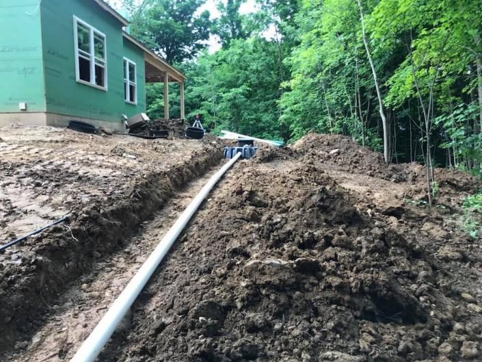 Mud-covered hillside beside a green house, with a long pipe running down the slope into a wooded area.