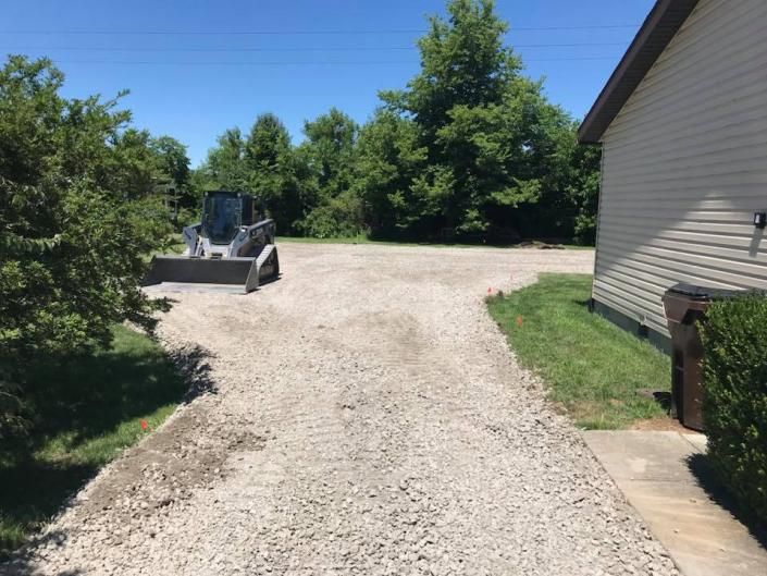 Gravel driveway beside a house, with a loader parked near trees under a blue sky.