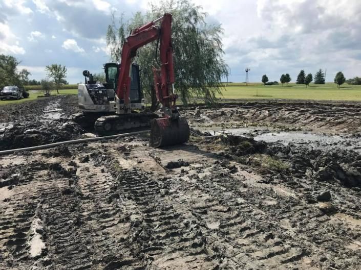Red excavator digging muddy ground in a flooded field under a cloudy sky