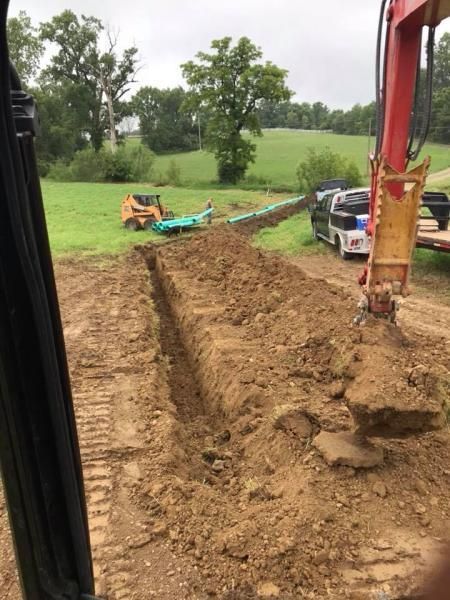Mud trench beside a red excavator in a rural field, with a small loader and pickup nearby.