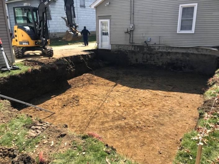 Excavated backyard beside a house, with a small backhoe and worker near the edge of a large dirt pit.
