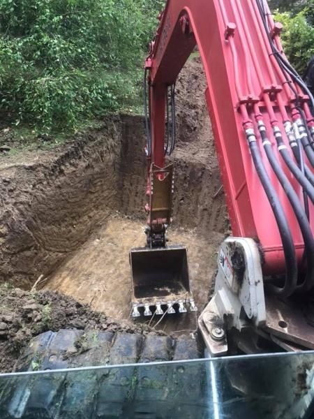 Red excavator digging a trench beside a wooded hillside