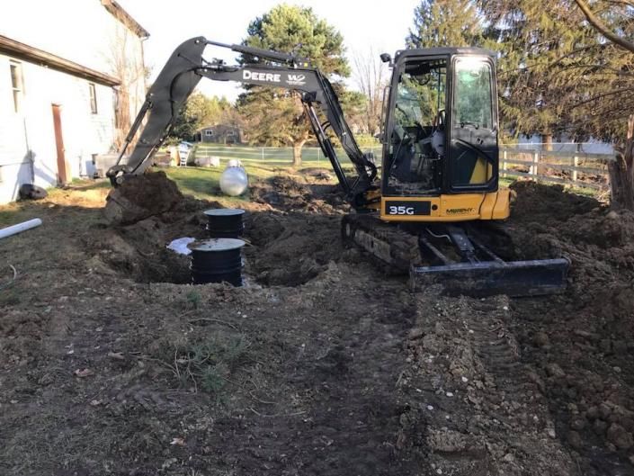 Yellow excavator digging in a muddy yard beside a house with trees in the background