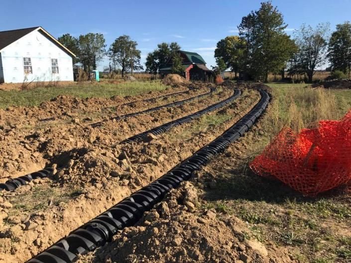 Black corrugated pipe laid through a dirt field beside a white house and trees, with orange safety netting nearby