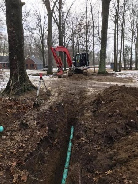 Red excavator digging a trench in a wooded yard, with a green pipe running through the ground.