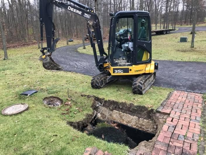 Mini excavator beside a large sinkhole in a grassy yard near a paved driveway