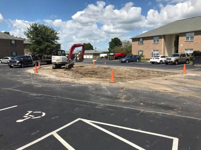 Road construction site with excavator, orange cones, and parked vehicles beside an apartment building.