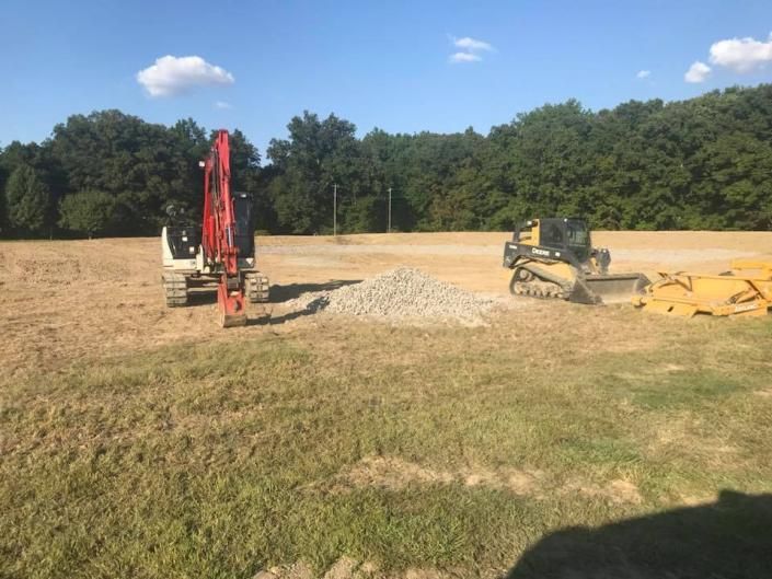 Construction equipment on a dirt field beside a gravel pile, with trees and blue sky in the background.