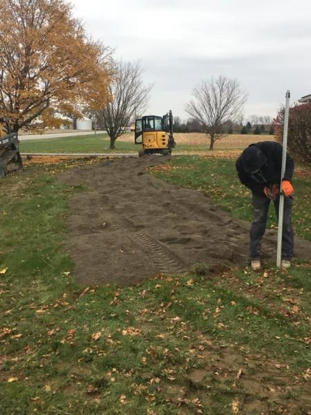 Workers preparing a dirt path beside a field, with a small yellow excavator in the background.