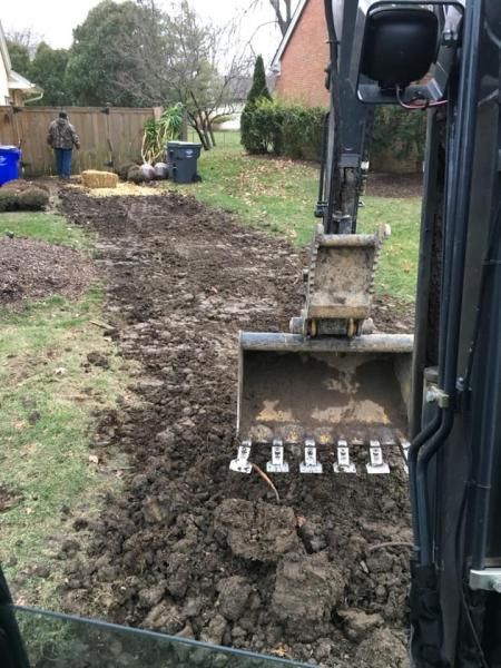 Excavator bucket digging muddy soil along a backyard path with a fence and bins in the background