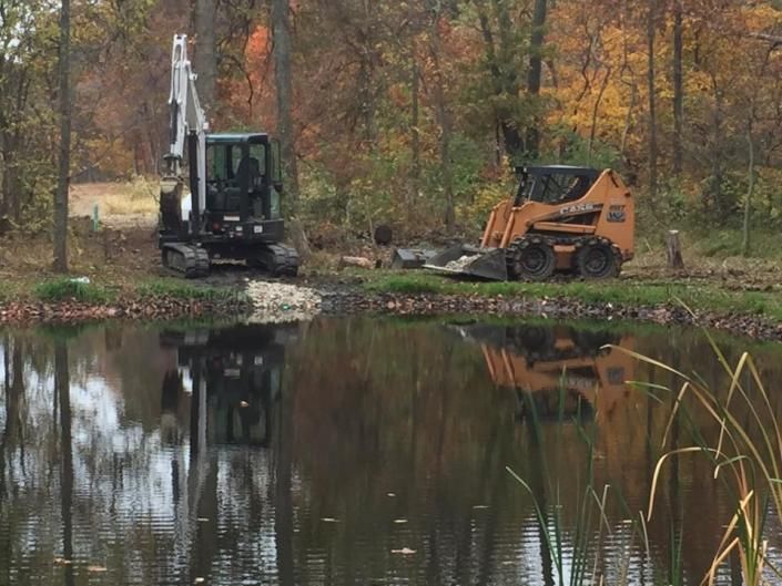 Excavator and skid steer by a pond with autumn trees reflected in the water