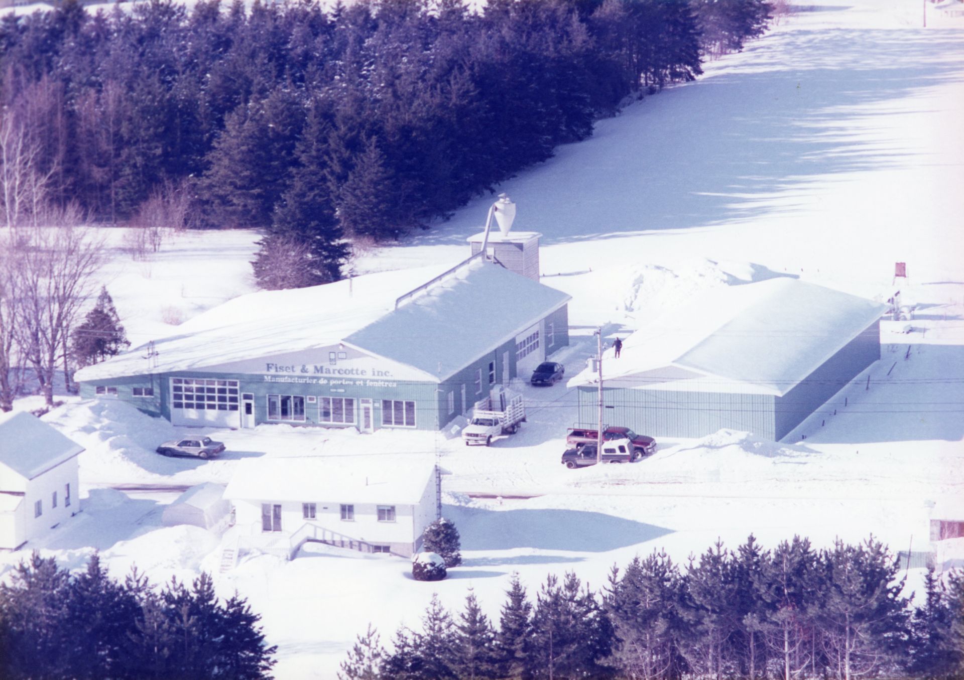 Ensemble de bâtiments enneigés aux toits bleu clair. Il comprend un grand hangar, de petites maisons et une tour de télécommunications, le tout entouré d'arbres.
