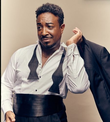 Man in formal wear, holding a jacket, looking to the side with a slight smile.