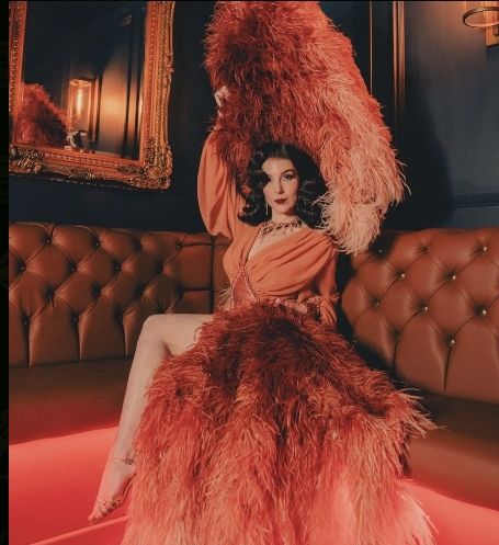 Woman in orange dress and feathers poses on a tufted leather couch.  Setting is a dimly lit room with a mirror.