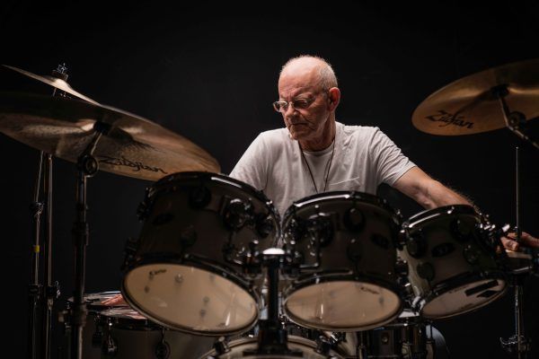 A drummer with glasses, wearing a white t-shirt, plays a drum set in front of a dark background.