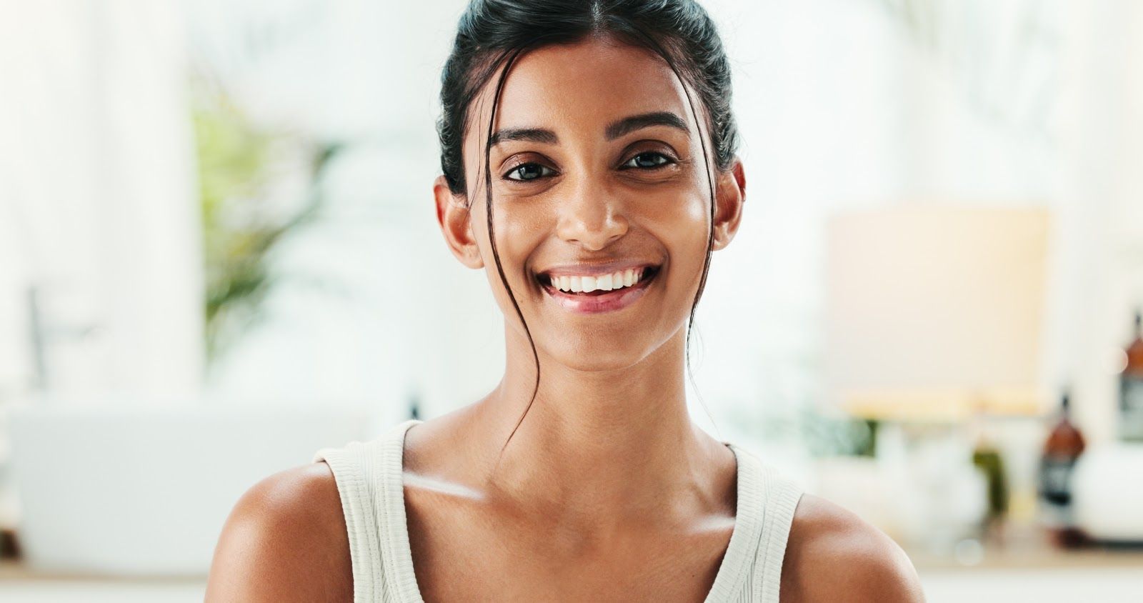 Smiling woman in a white tank top standing in a bright, softly blurred room