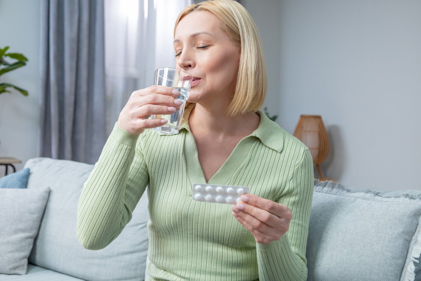 Woman sitting on a couch, drinking water and holding a blister pack of pills