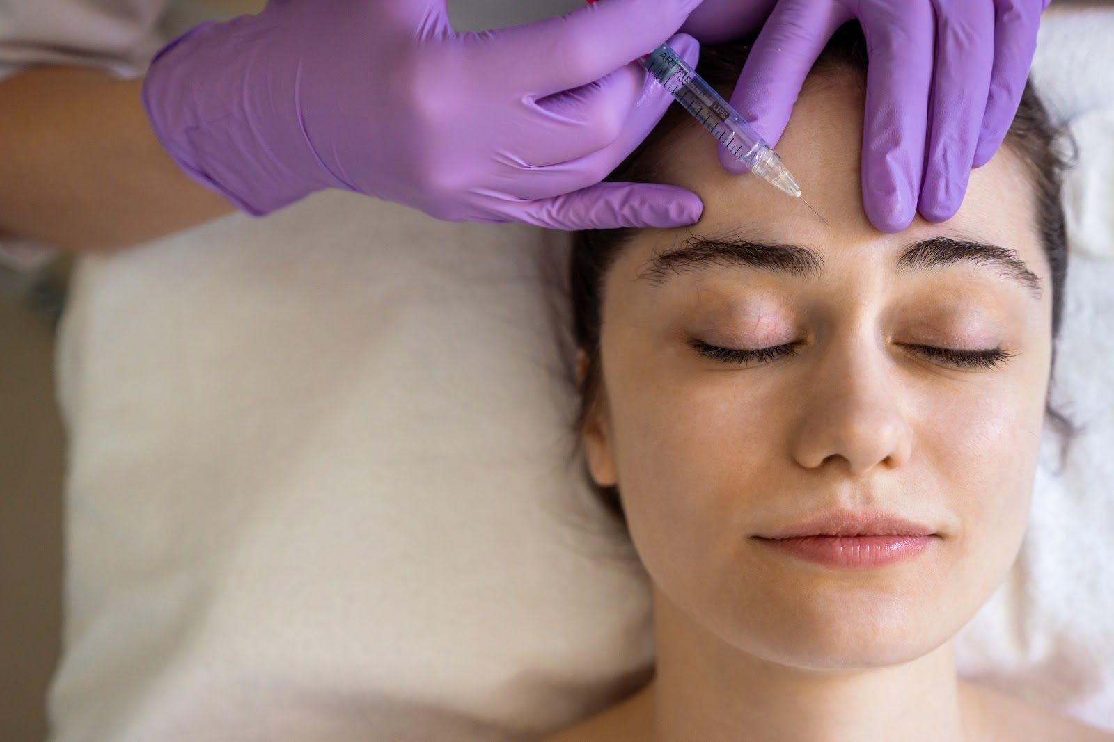 Gloved hands apply treatment to a woman’s forehead as she lies with eyes closed on a treatment bed