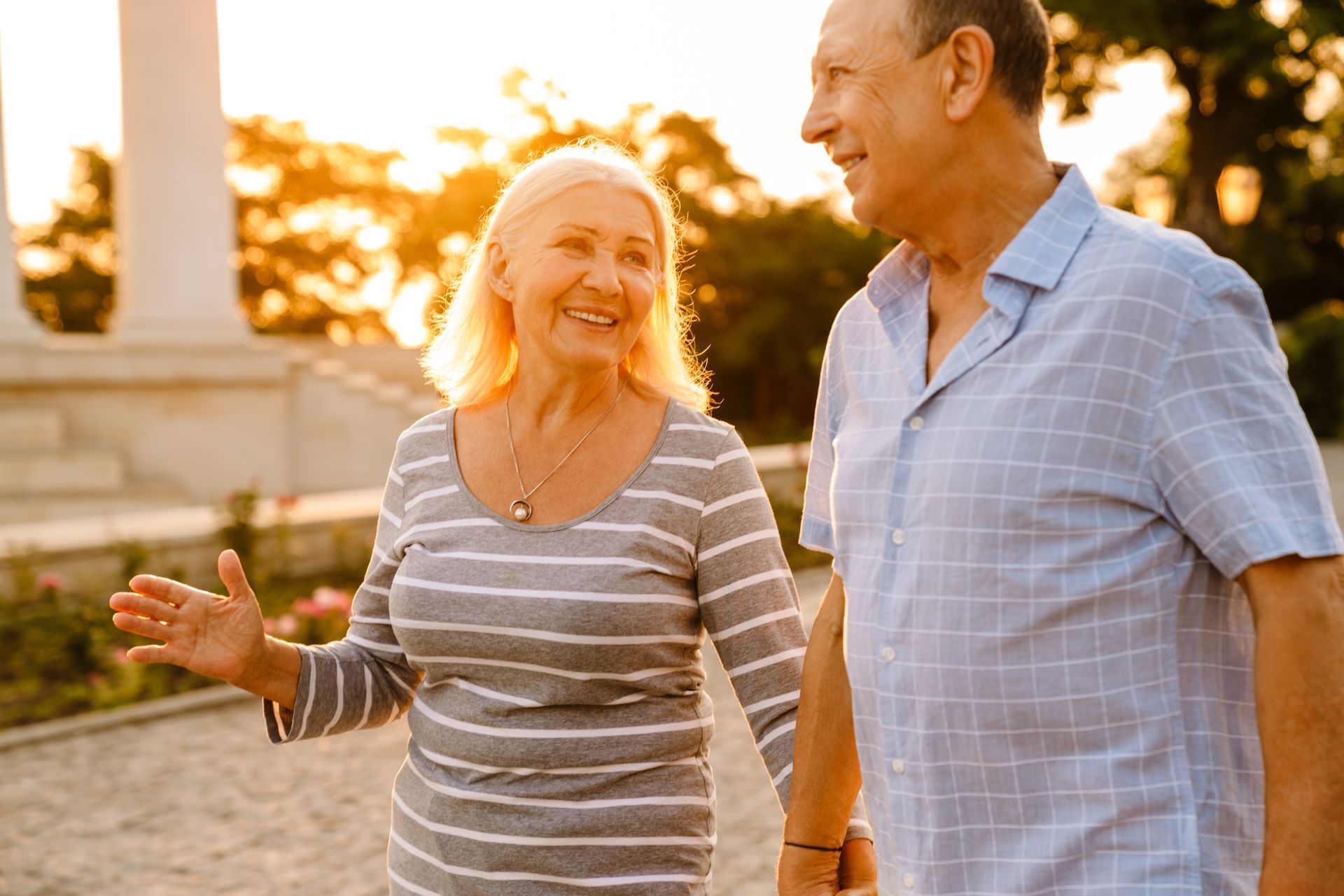 An elderly couple is walking in a park holding hands and smiling.