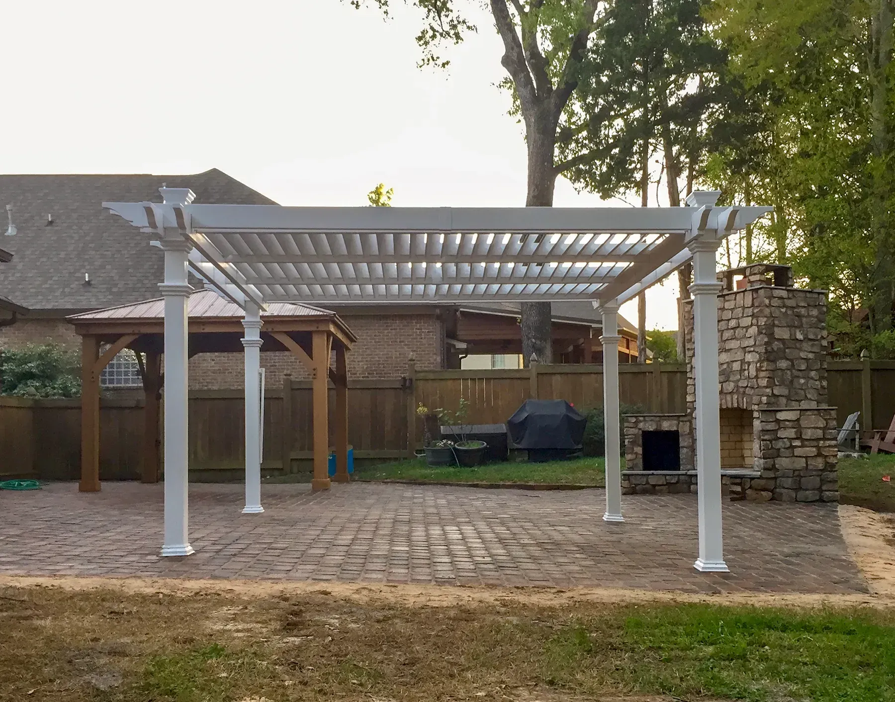 A White Pergola Is Sitting On Top Of A Brick Patio Next To A Stone Fireplace - Raymond, MS - Parker Roofing & Remodeling