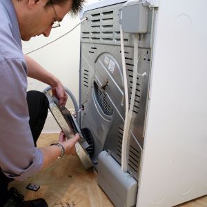 Man connecting a hose to the back of a white appliance, possibly a dryer, indoors.