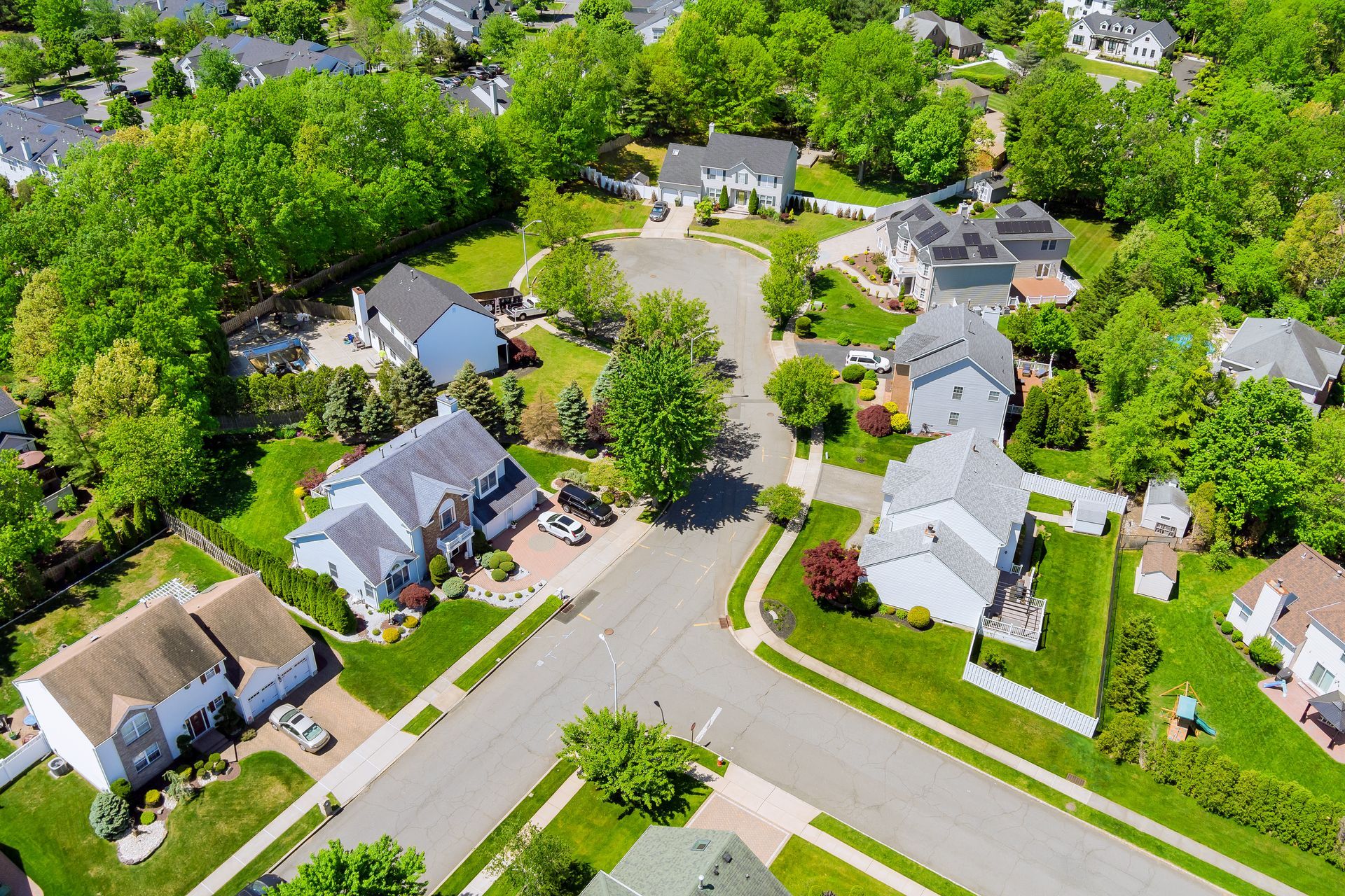 Aerial view of a suburban neighborhood with houses, green lawns, trees, and roads.