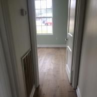 A view down a hallway with light wood flooring toward a bedroom doorway and a window looking out onto a residential street.
