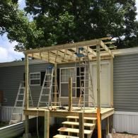 A wooden deck and pergola frame being constructed over the entrance of a grey mobile home, featuring two ladders.