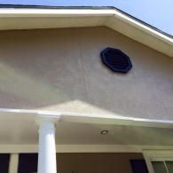 A tan house exterior with a white column supporting a porch roof and a dark octagonal attic vent on the gable wall.