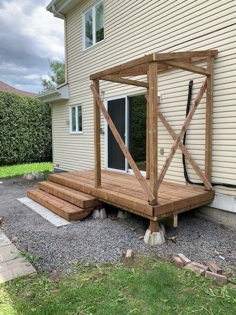 Une terrasse en bois adossée à l'arrière d'une maison beige, composée d'une structure à ossature bois et de deux marches menant au jardin.