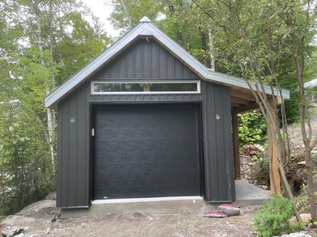 Un hangar aux parois sombres, avec une porte de garage centrale, une fenêtre rectangulaire étroite au-dessus et un porche latéral couvert.