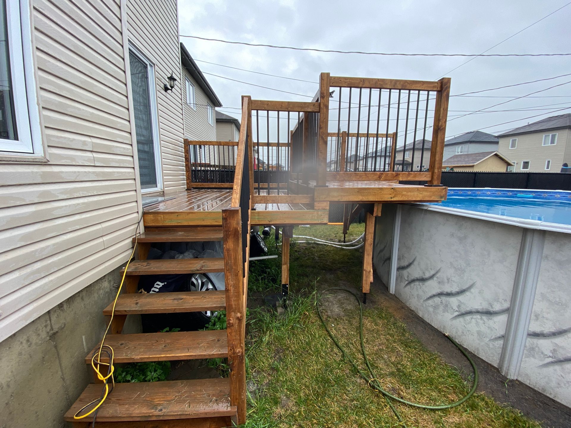 Une terrasse en bois avec escaliers et rampes menant &agrave; une piscine hors-sol, situ&eacute;e entre des maisons r&eacute;sidentielles